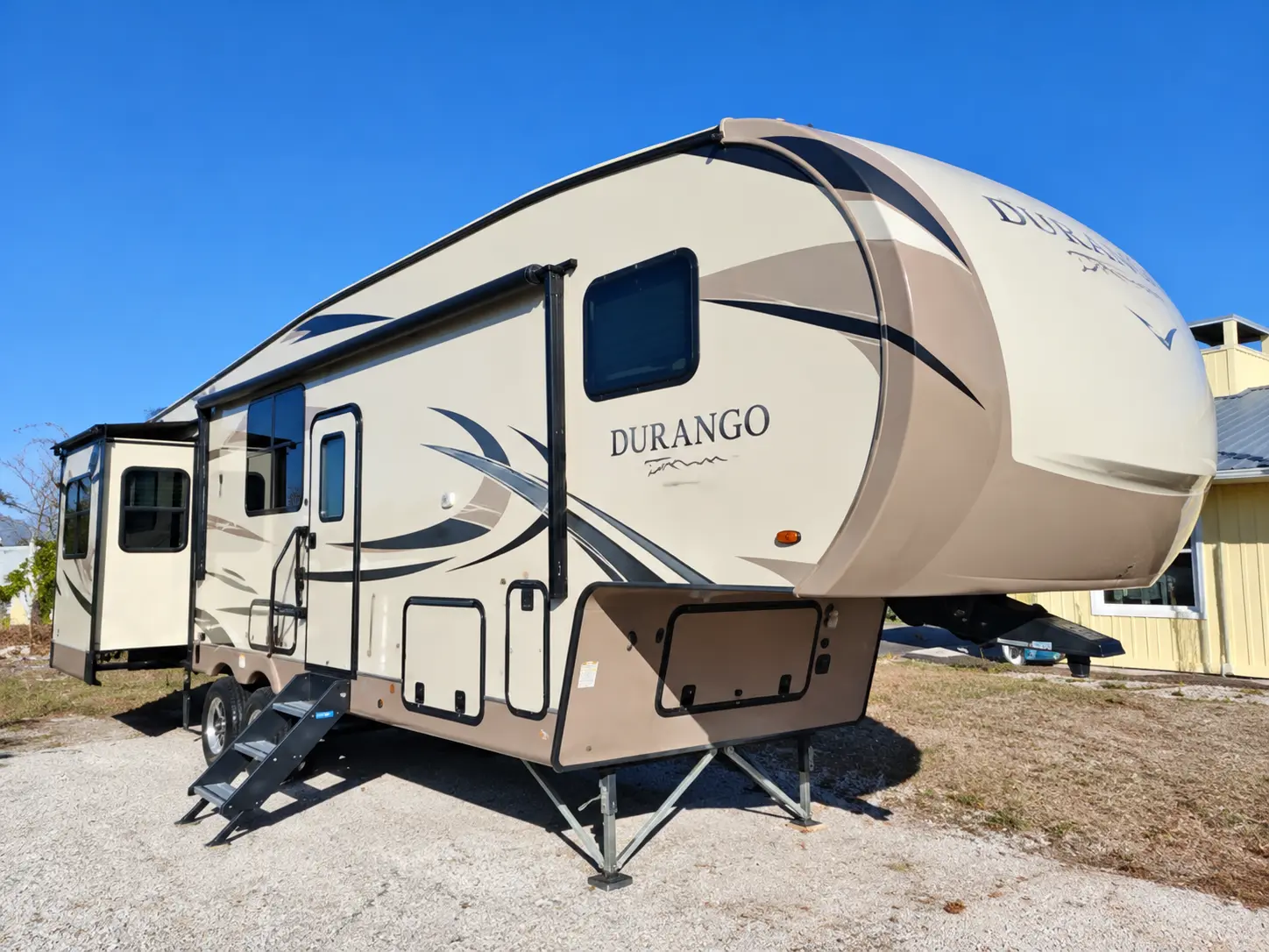 Cream and tan Durango travel trailer with slide-out extension parked on gravel lot under clear blue sky
