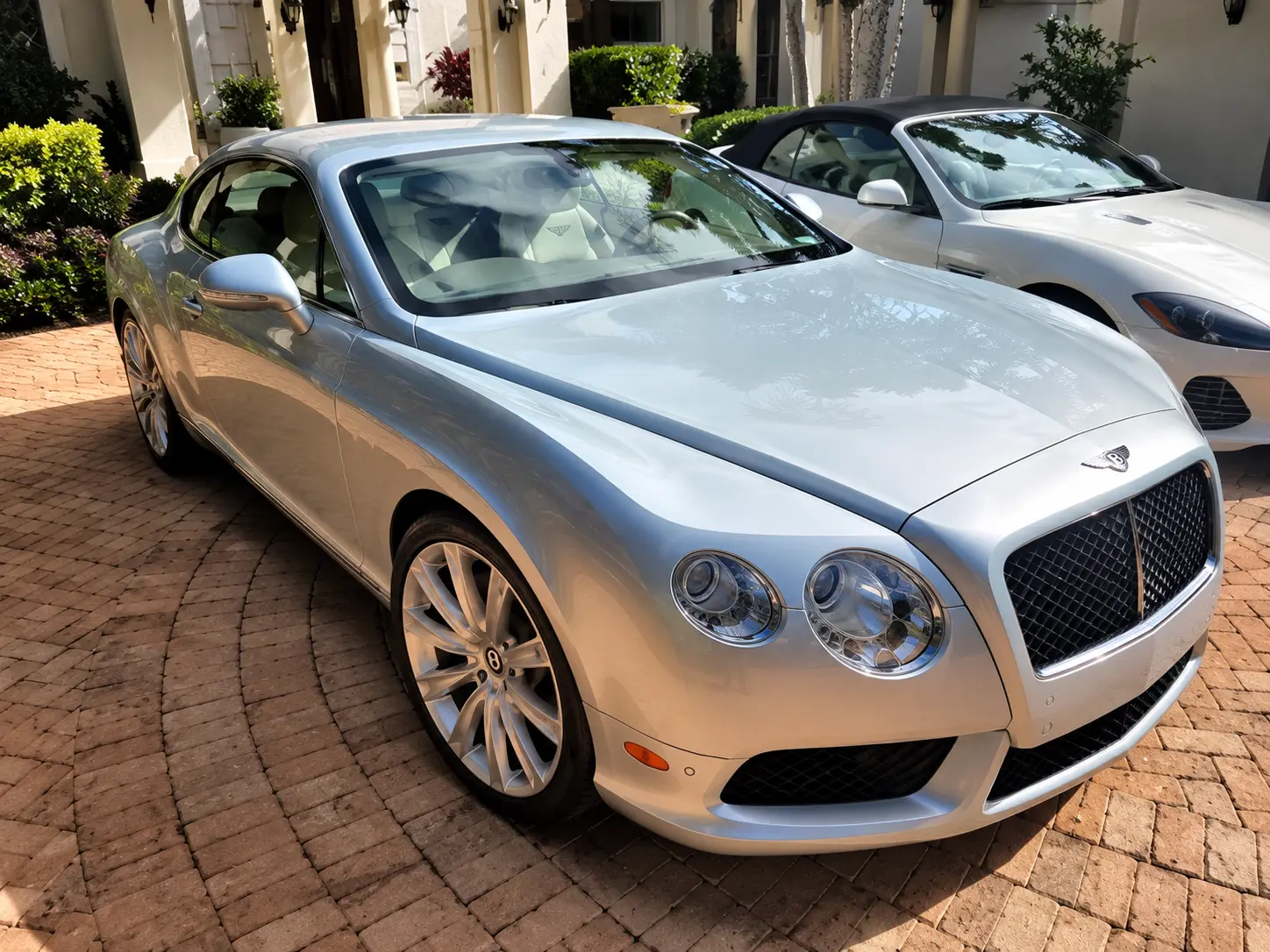 White Bentley Continental GT coupe parked in brick driveway with white residential buildings and landscaping in background